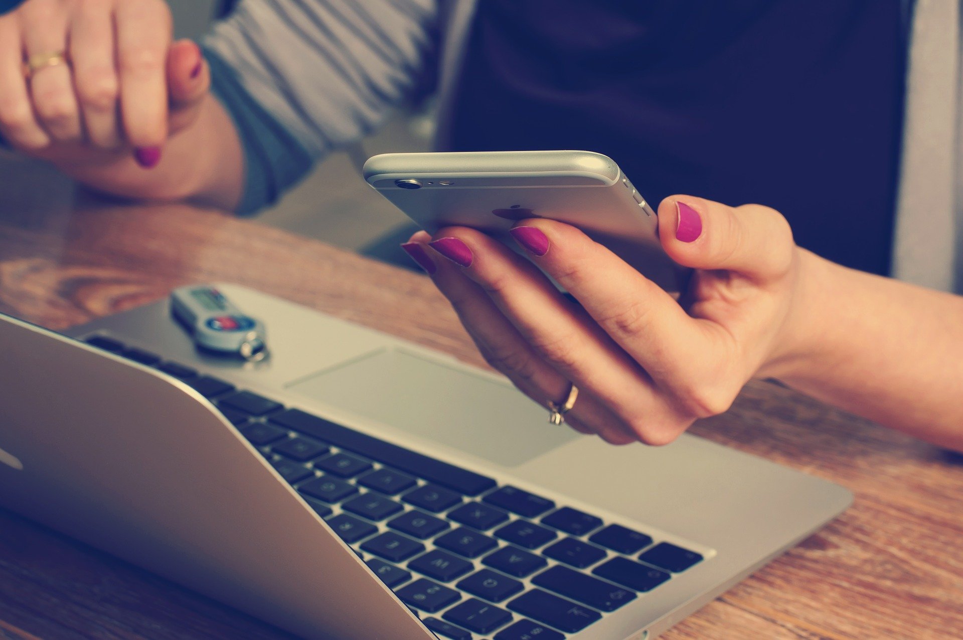 Hand with painted nails holding a smartphone, with a laptop on the table in front