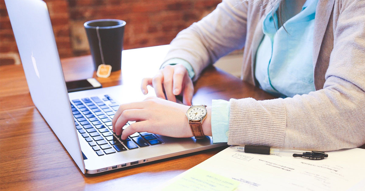 Woman working on a laptop on a wooden table, with papers and mugs around it