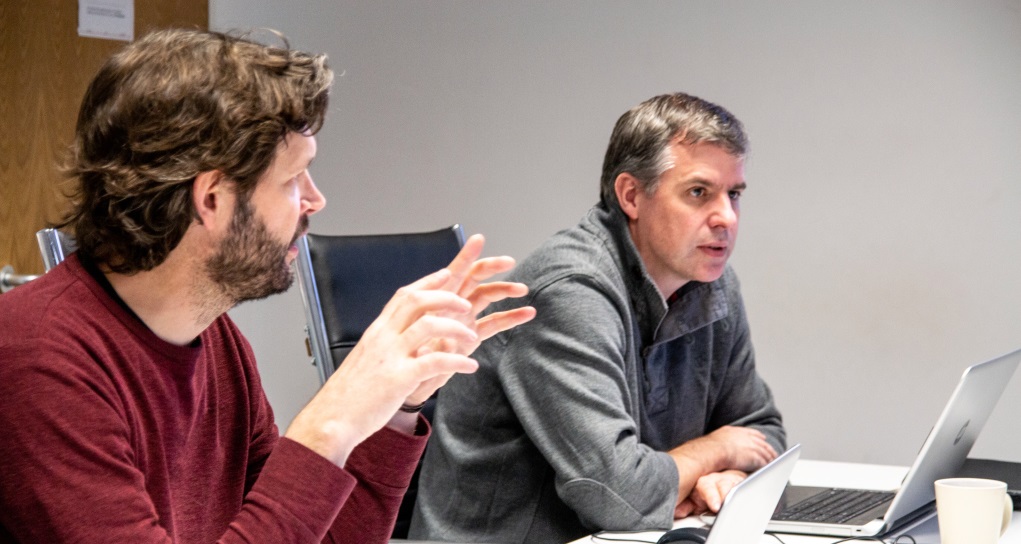 Two men having a discussion at a table, one with a laptop