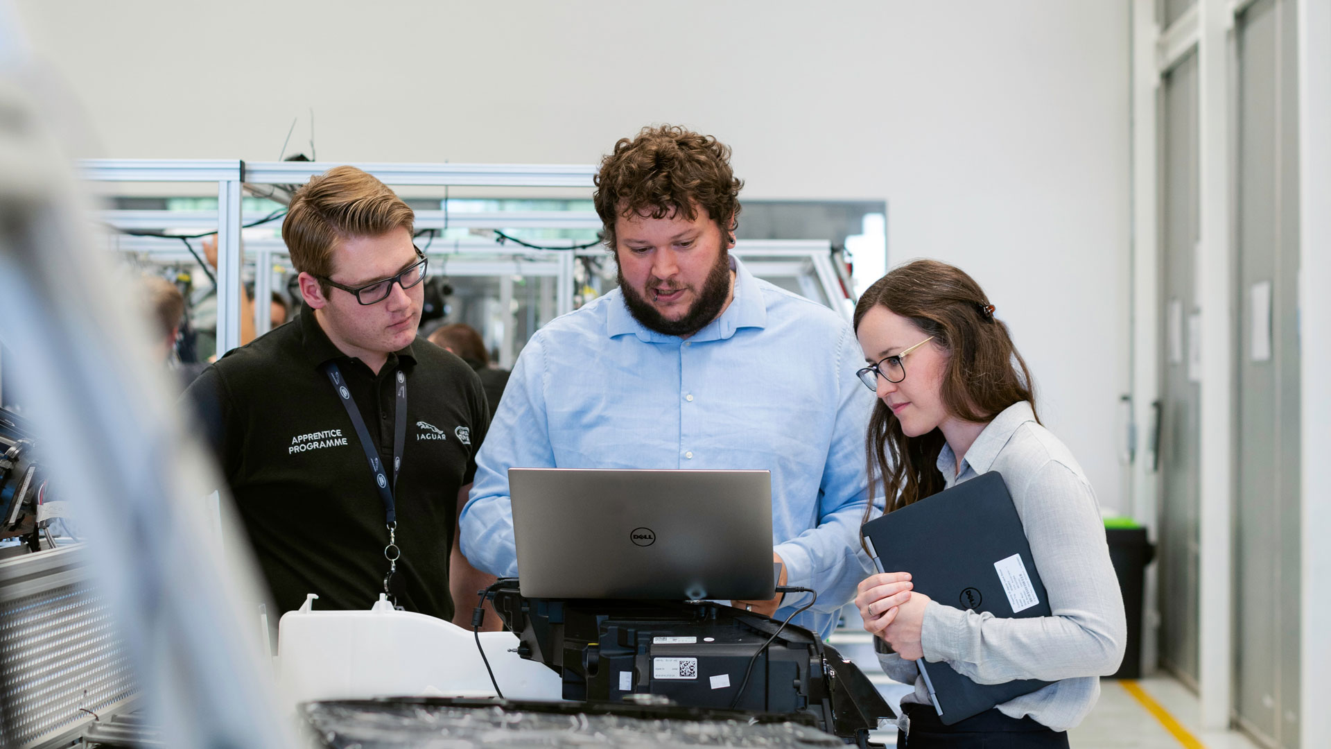 Three people in factory environment, looking at a laptop screen