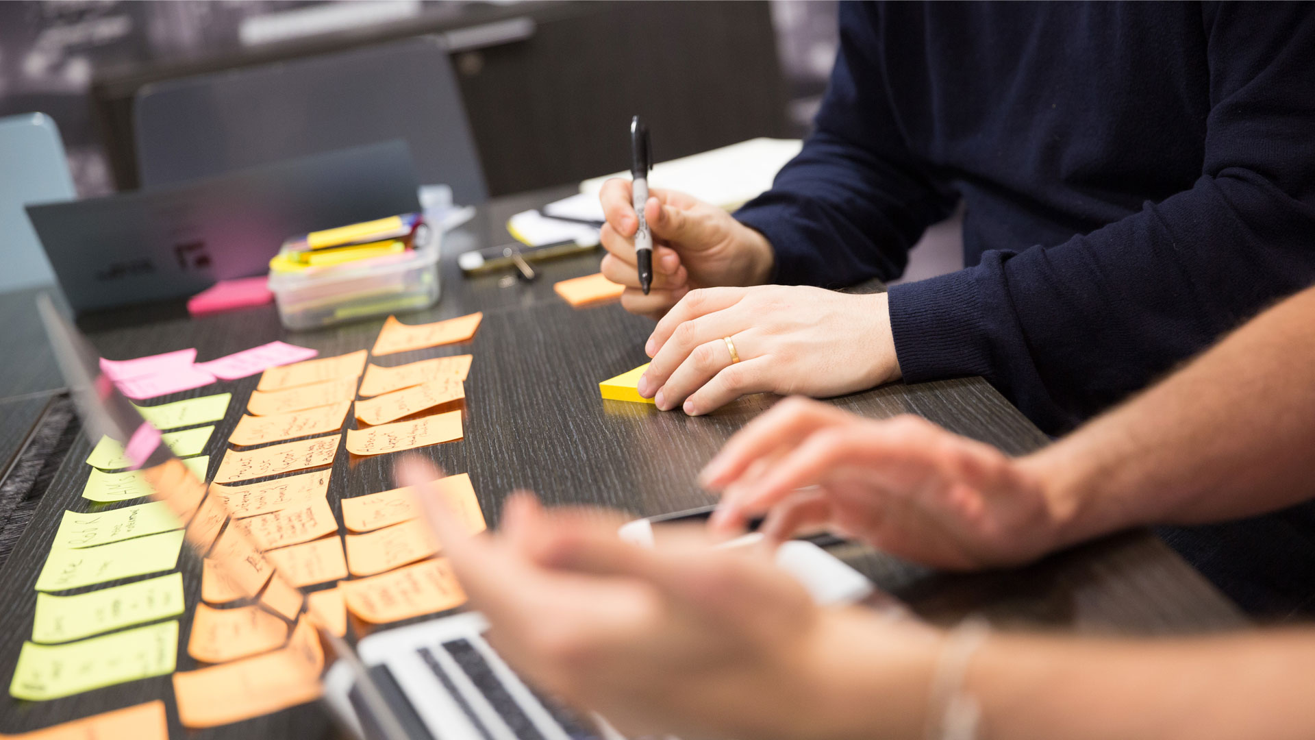 Two people writing on post-it notes with a laptop in the foreground