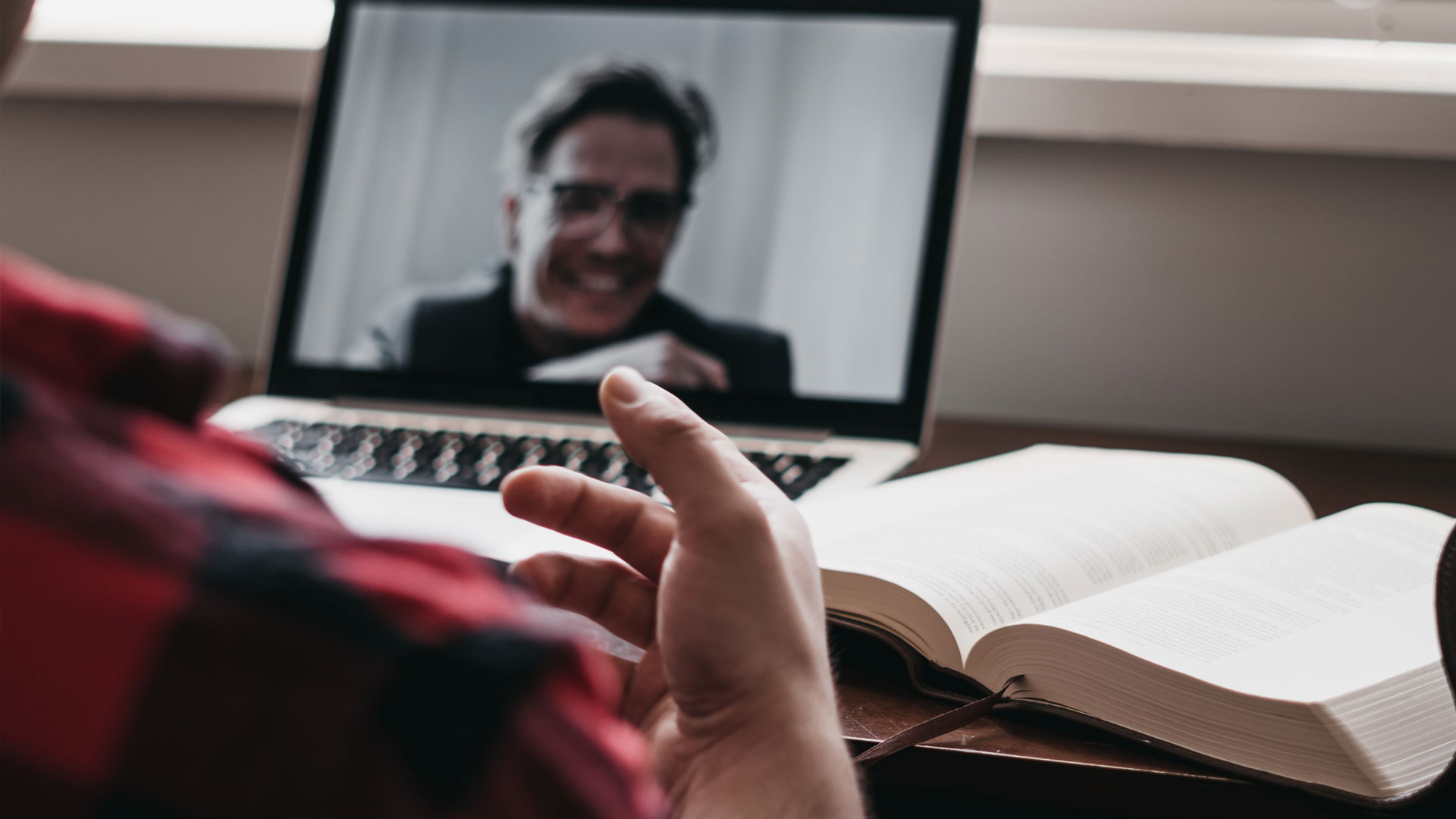 Laptop showing man on video call, with book and other person in foreground