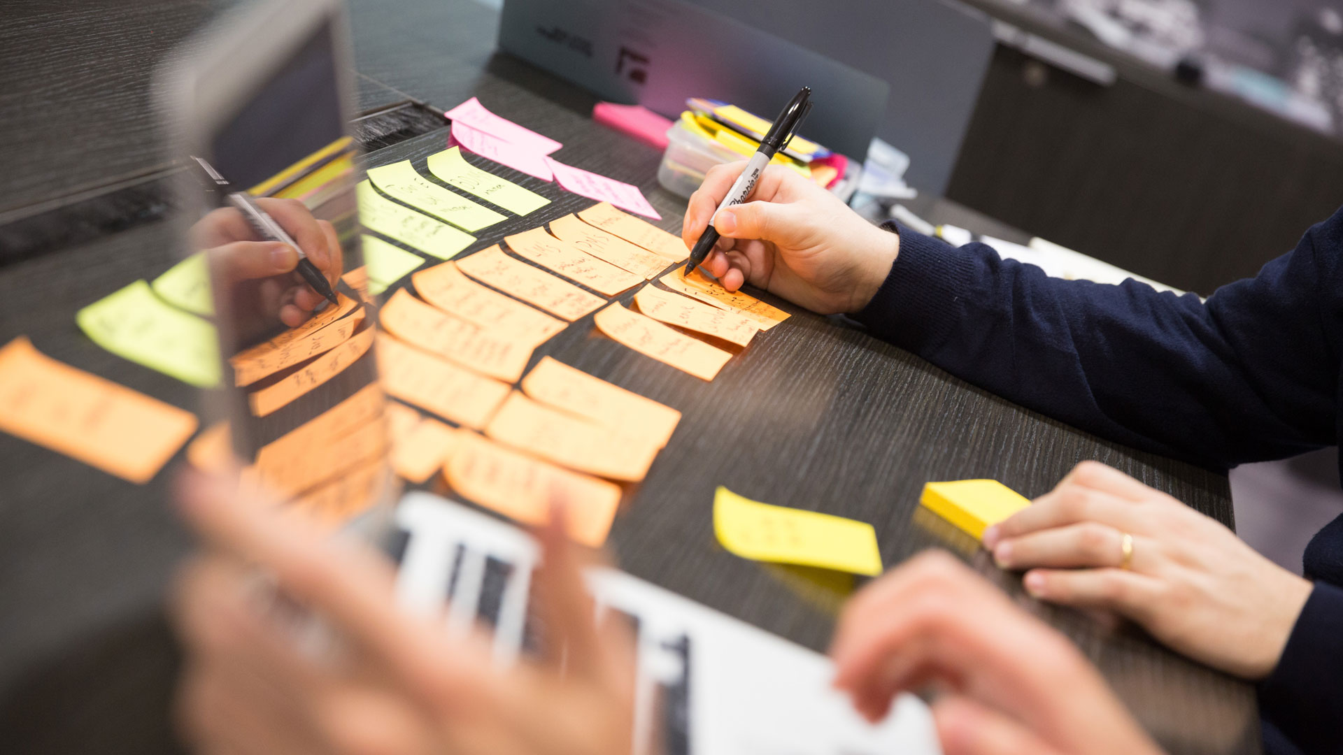 Table covered in post-it notes, with person writing on post-its and someone working on a laptop in foreground