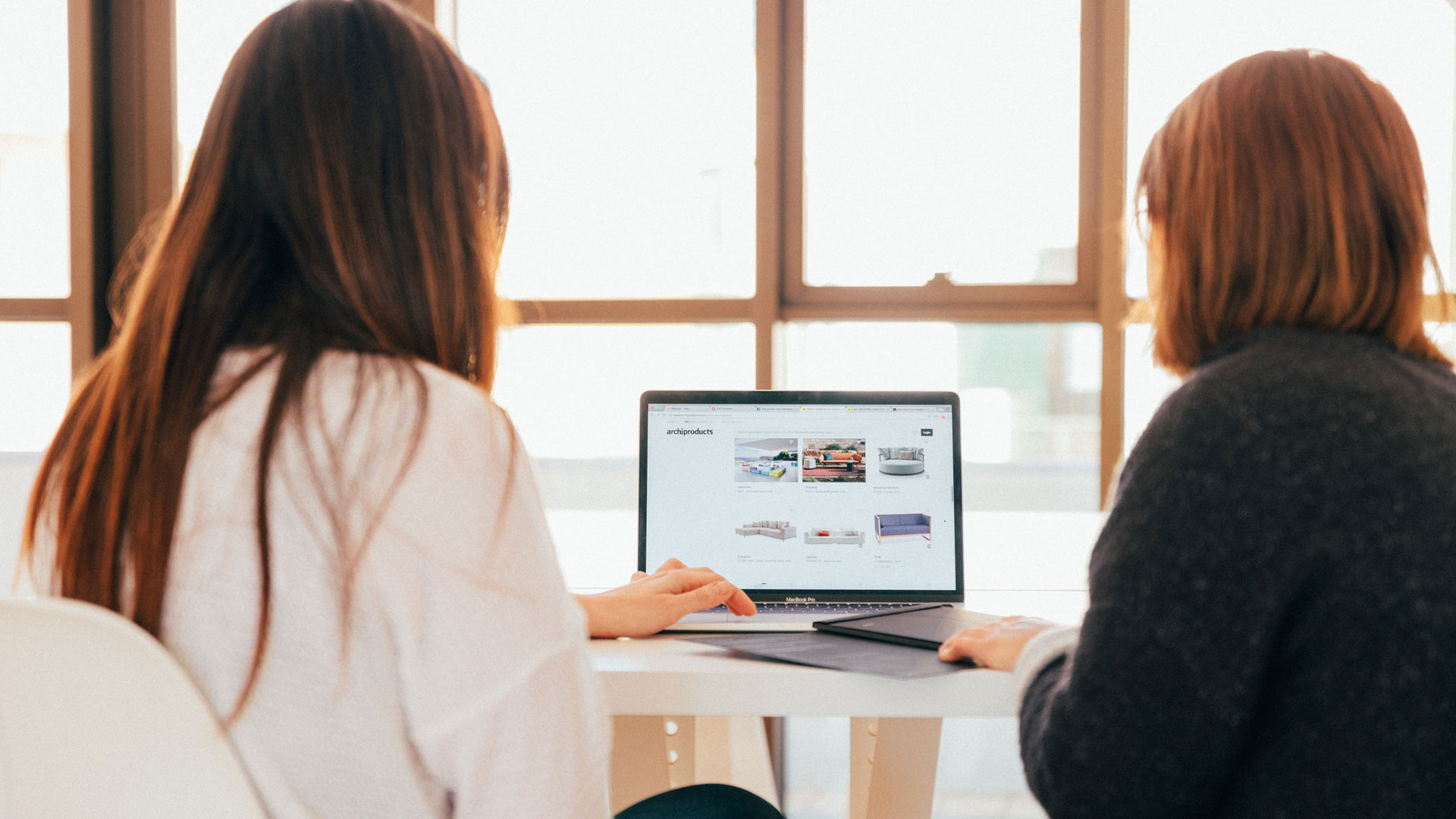 Two people working on a laptop in front of large windows