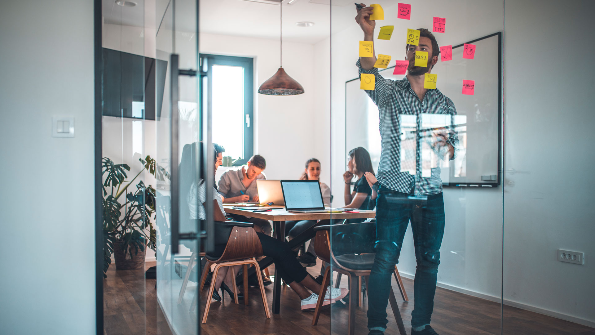 Meeting room with people at a table and one person sticking post-it notes on a glass wall